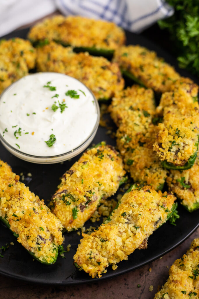 Air fryer jalapeno poppers on a large black matte plate surrounding a small bowl of ranch sauce.