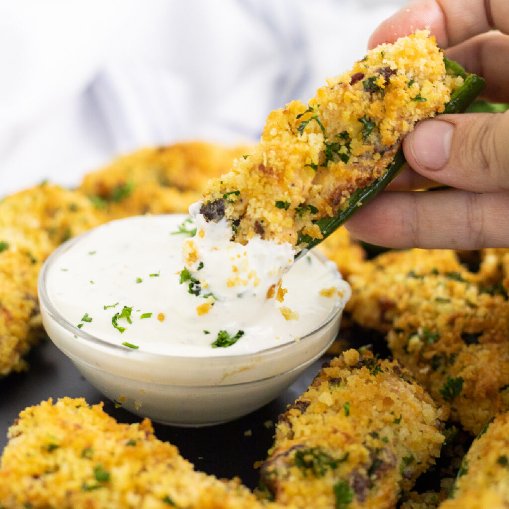 An air fryer jalapeno popper being dipped into a small glass bowl of ranch dressing surrounded by more poppers.