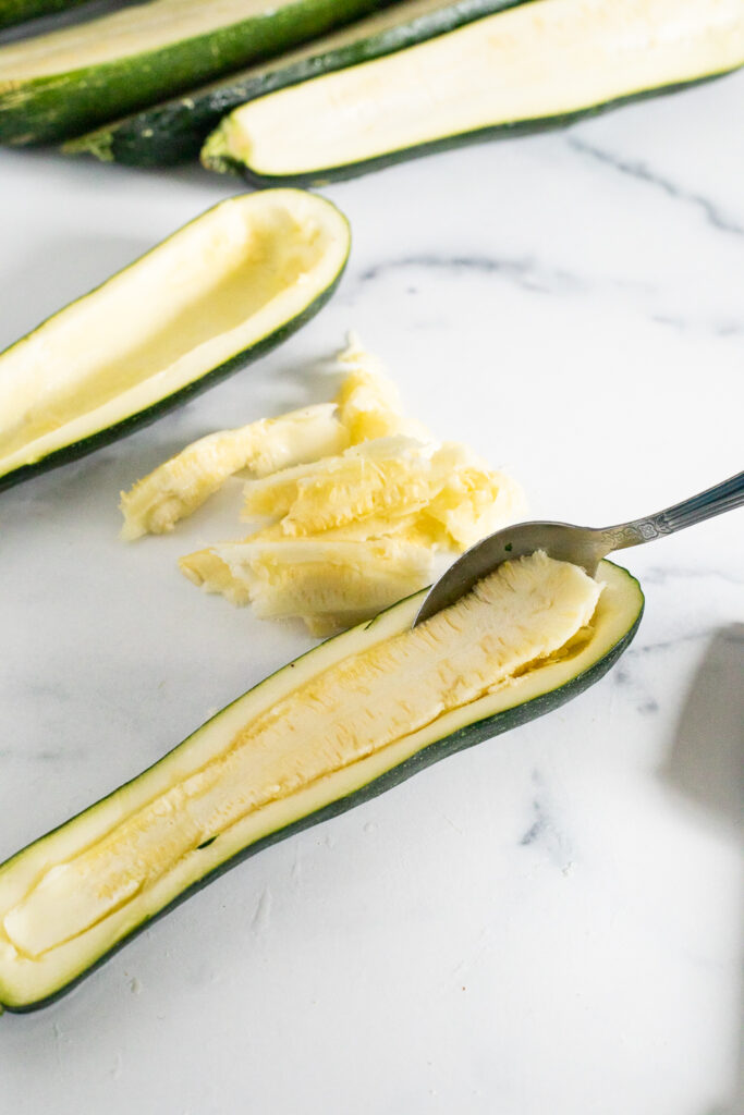 A spoon hollowing out the center of the zucchini on a white marble counter top to prepare it to be stuffed. 