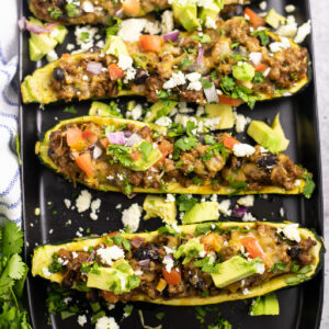 Overhead view of 3 taco zucchini boats on a black matte plate topped with fresh avocado, diced red onion, chopped tomatoes, and crumbled queso fresco.
