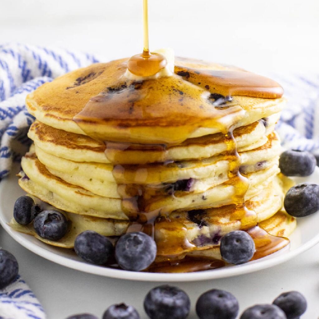 Syrup being drizzled and running down a stack of buttermilk blueberry pancakes on a small plate surrounded by fresh blueberries.