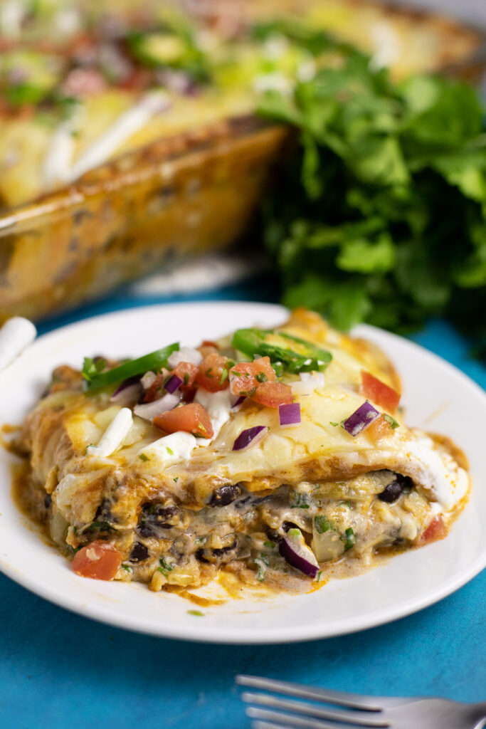 A piece of black bean enchilada casserole on a small white plate with a casserole dish and a bunch of fresh cilantro behind.