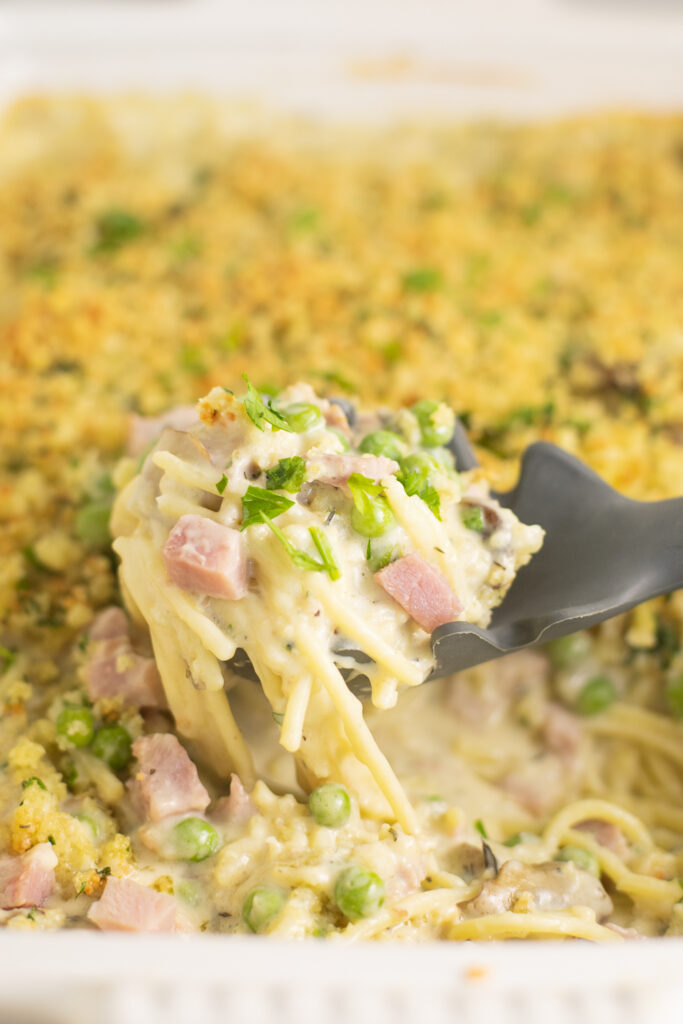 Close up of a silicone spoon scooping a big scoop of ham tetrazzini with diced ham and peas topped with a crispy topping out of a large white casserole dish.