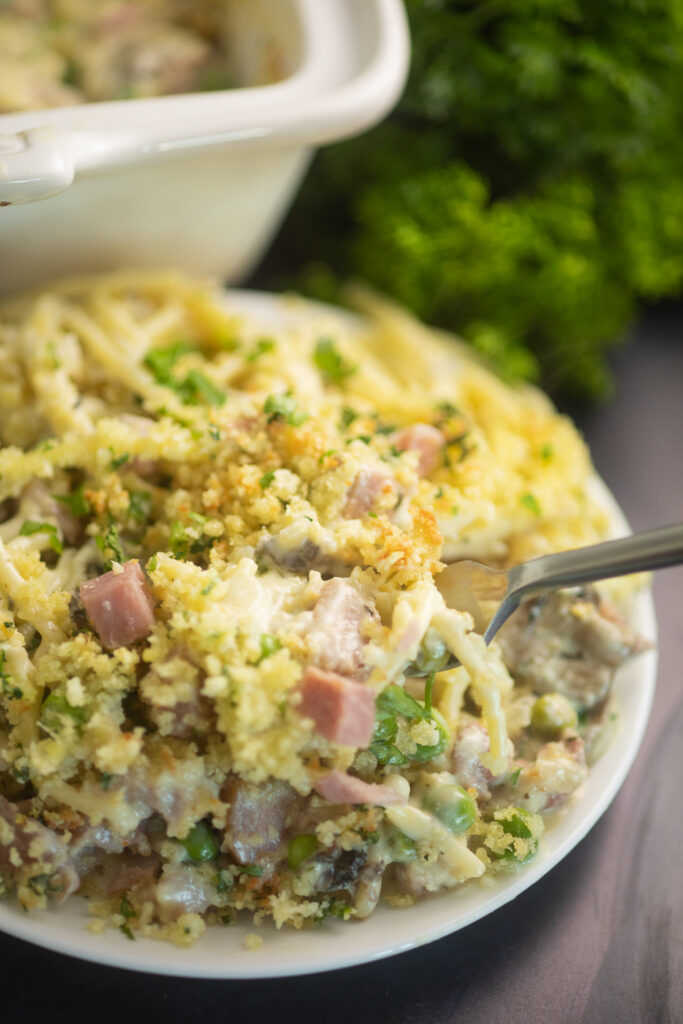 Ham tetrazzini on a small white plate with a fork in it with a large white casserole dish and a bunch of parsley