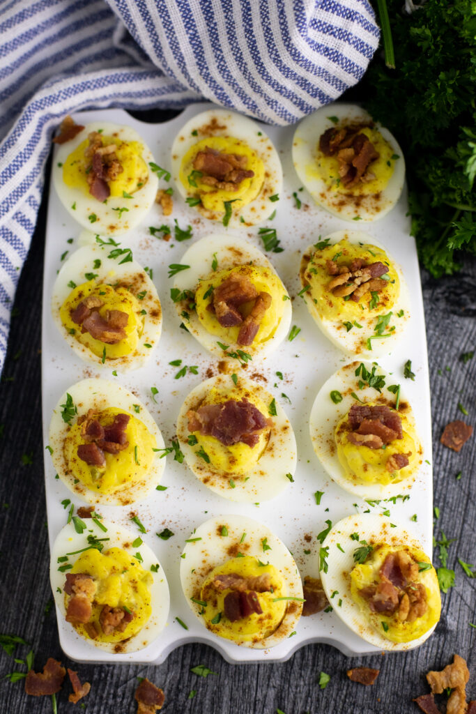Overhead view of a dozen horseradish deviled eggs topped with crumbled bacon and fresh chopped parsley on a rustic wood background next to a striped linen and a bunch of fresh parsley.