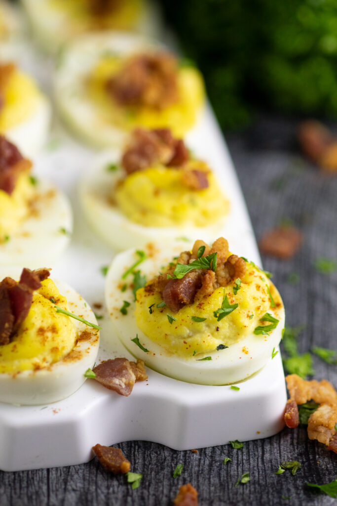 Side view of a horseradish deviled egg topped with crumbled bacon and fresh chopped parsley on a serving tray with a rustic wood background.