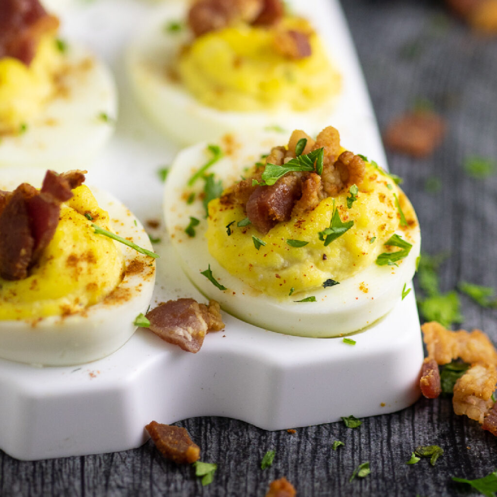 Side view of a horseradish deviled egg topped with crumbled bacon and fresh chopped parsley on a serving tray with a rustic wood background.