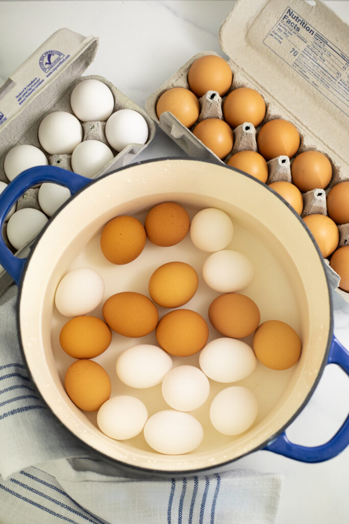 An overhead of a ceramic pot with 18 eggs, half white and half brown, covered with water with 2 cartons of eggs behind and a linen napkin next to the pot.