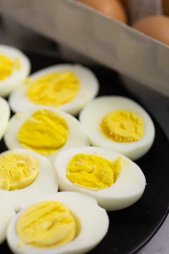 Hard boiled egg halves on a black plate with a carton of brown eggs behind.