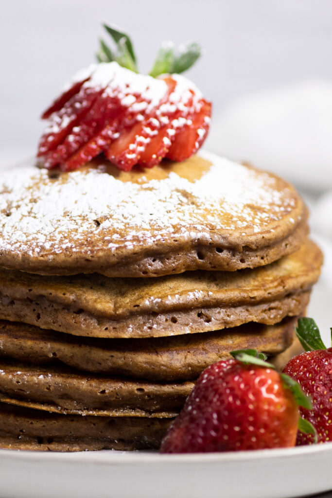 Close up side view of a stack of chocolate pancakes topped with a sliced strawberry and a dusting of powdered sugar
