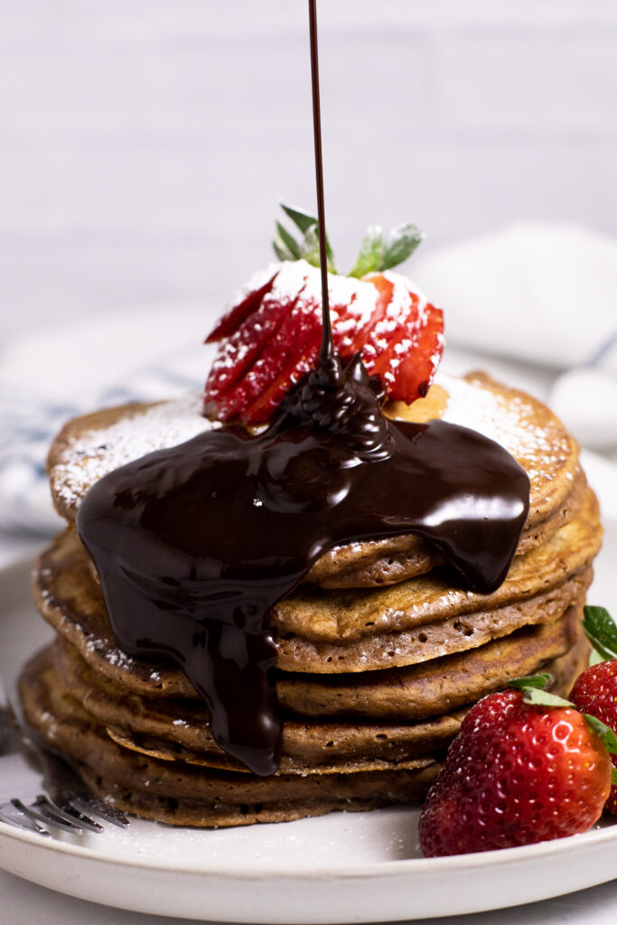 A stack of chocolate pancakes topped with a sliced strawberry and chocolate sauce being drizzled on top on a small white plate surrounded by strawberries