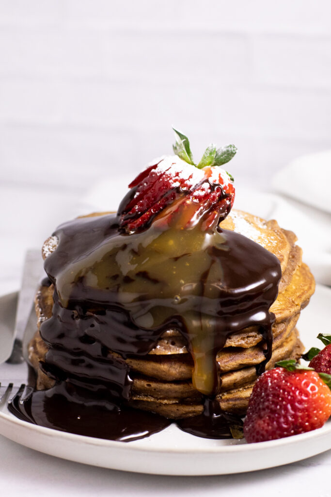 Stack of chocolate pancakes topped with a sliced strawberry and chocolate and caramel sauce dripping down.