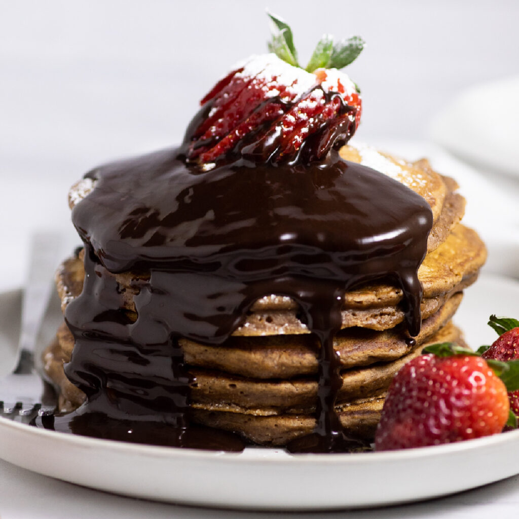 Stack of chocolate pancakes on a white plate topped with a sliced strawberry and chocolate sauce running down