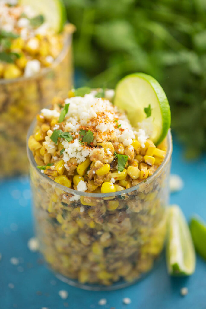 Esquites in a small glass cup topped with crumbled cotija cheese, fresh cilantro, and a lime slice with a