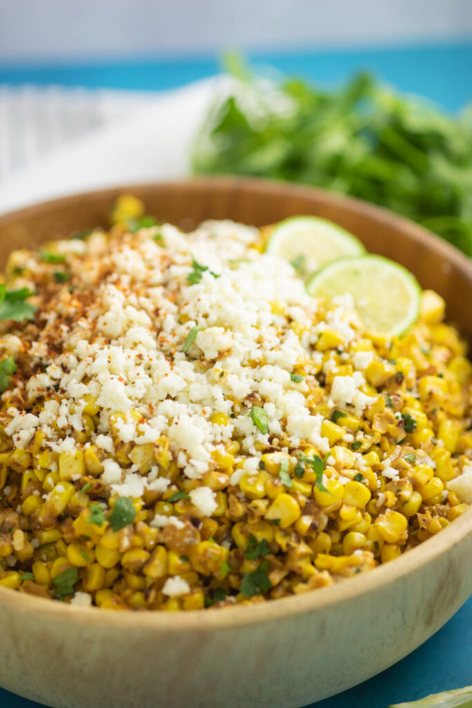 Esquites in a large wooden bowl topped with crumbled cotija cheese, fresh cilantro, and two lime slices with a bunch of cilantro and a white linen in the background.