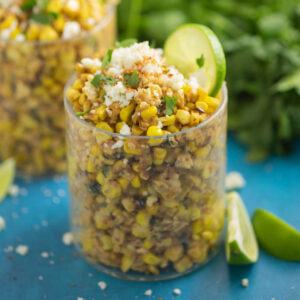 Esquites in a small glass cup topped with crumbled cotija cheese, fresh cilantro, and a lime slice with a bunch of cilantro and another esquites cup in the background.