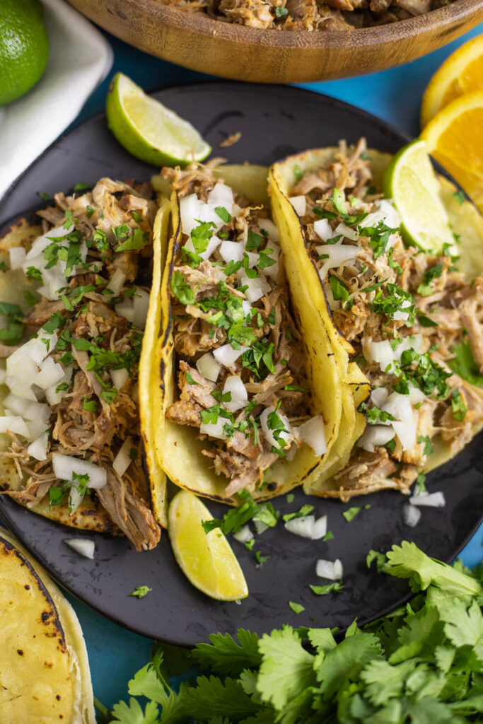 Overhead view of three instant pot carnitas tacos topped with onions and cilantro next to lime wedges, a bunch of fresh parsley and a bowl with instant pot carnitas behind.