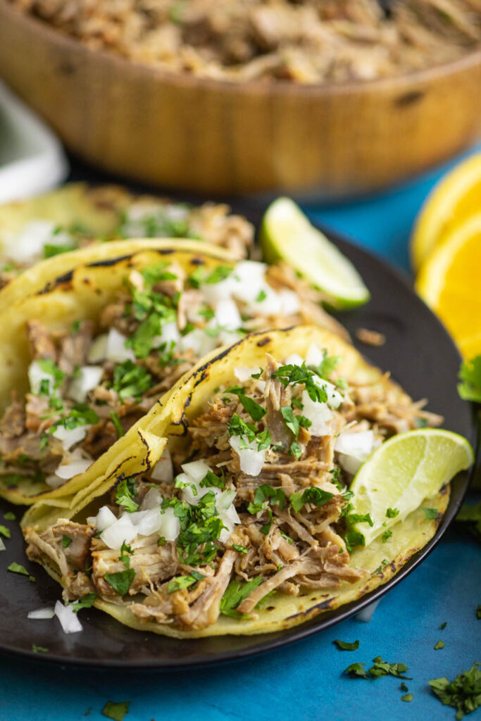 Instant Carnitas taco topped with chopped onions and fresh cilantro next to a lime wedge on a plate with two other tacos behind.