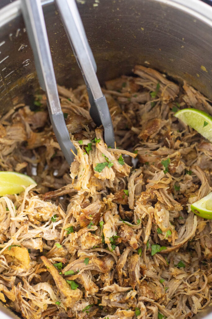 Crispy carnitas in an instant pot with lime wedges and cilantro and tongs pulling some out.