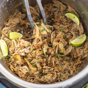 Crispy carnitas in an instant pot with lime wedges and cilantro and tongs pulling some out.