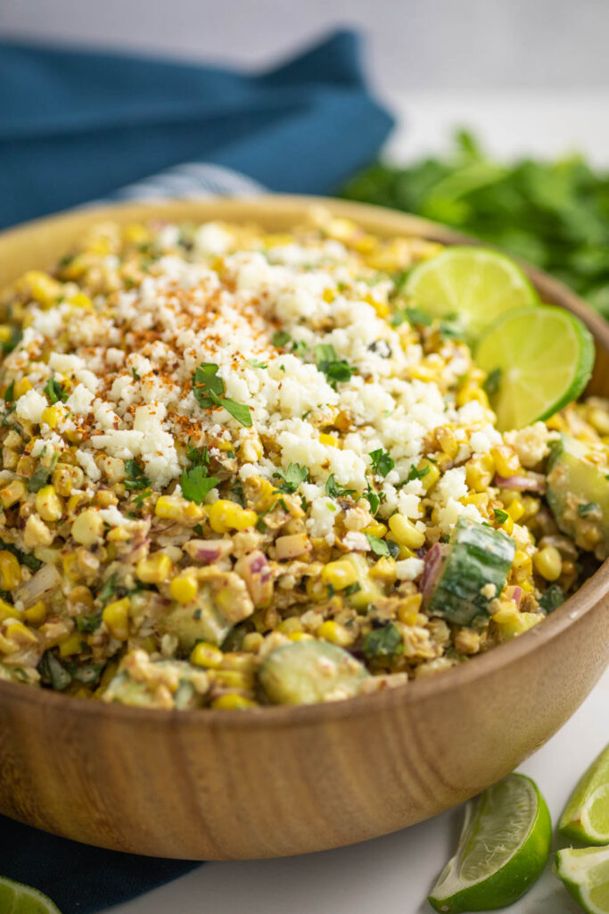 Mexican street corn salad in a wooden bowl topped with lime slices, cotija cheese, and chile lime seasoning with a teal linen napkin and bunch of fresh cilantro behind the bowl.