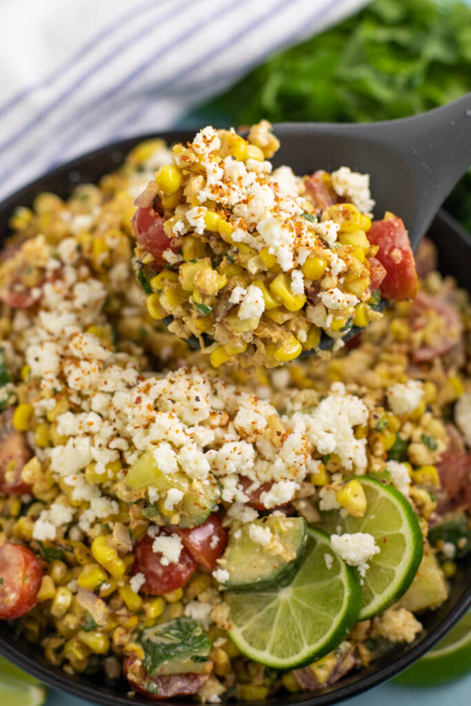 Silicone spoon with a large scoop of Mexican street corn salad with cherry tomatoes added. Spoon is hovering over a skillet full of the same salad with fresh lime slices
