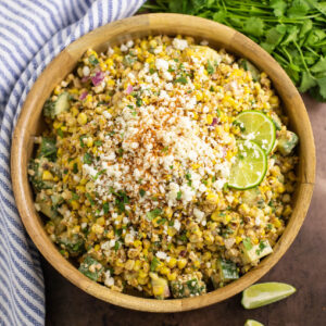 Overhead view of Mexican street corn salad in a large wooden bowl topped with cotija cheese, chile lime seasoning, and lime slices next to a bunch of fresh cilantro and a striped linen napkin on a rustic background.