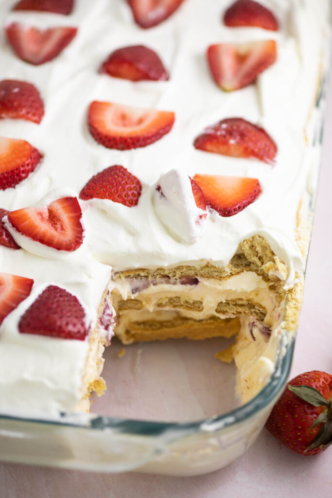 Overhead view of a strawberry icebox cake topped with whipped cream and sliced strawberries with a piece missing so the inside layers are visible.