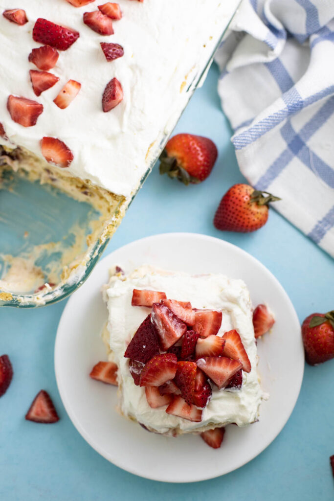 Overhead view of a slice of strawberry icebox cake on a small white plate next to the full cake in a glass pan surrounded by fresh strawberries and a plaid linen