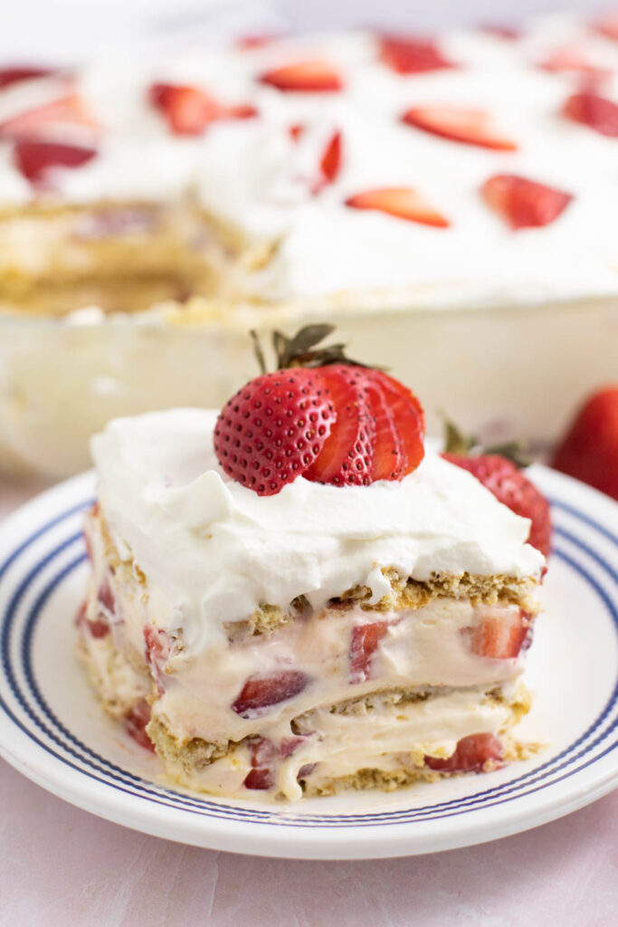 Square slice of strawberry icebox cake topped with a fresh strawberry on a small white plate with the rest of the cake in a glass dish behind