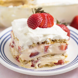 Square slice of strawberry icebox cake topped with a fresh strawberry on a small white plate with the rest of the cake in a glass dish behind