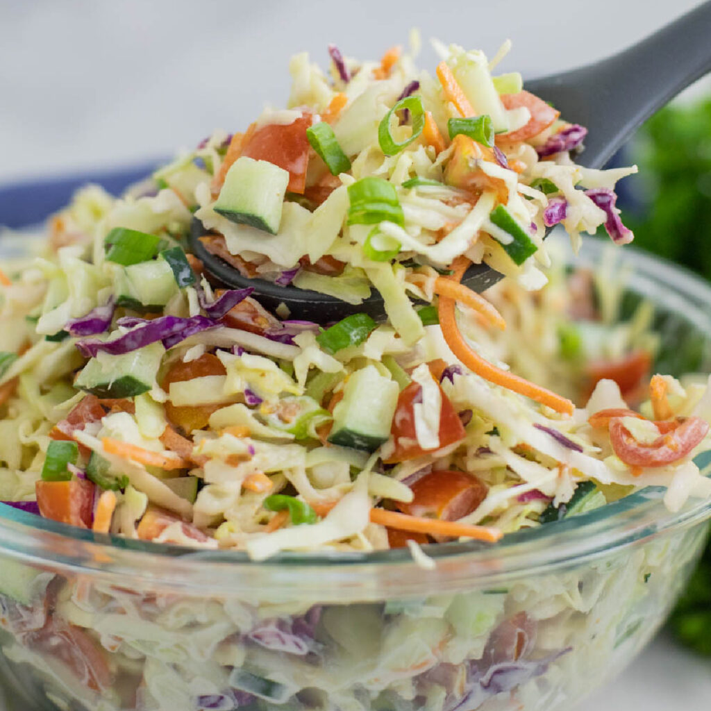Summer slaw with a creamy tangy dressing in a glass bowl with a silicone spoon taking a scoop out.