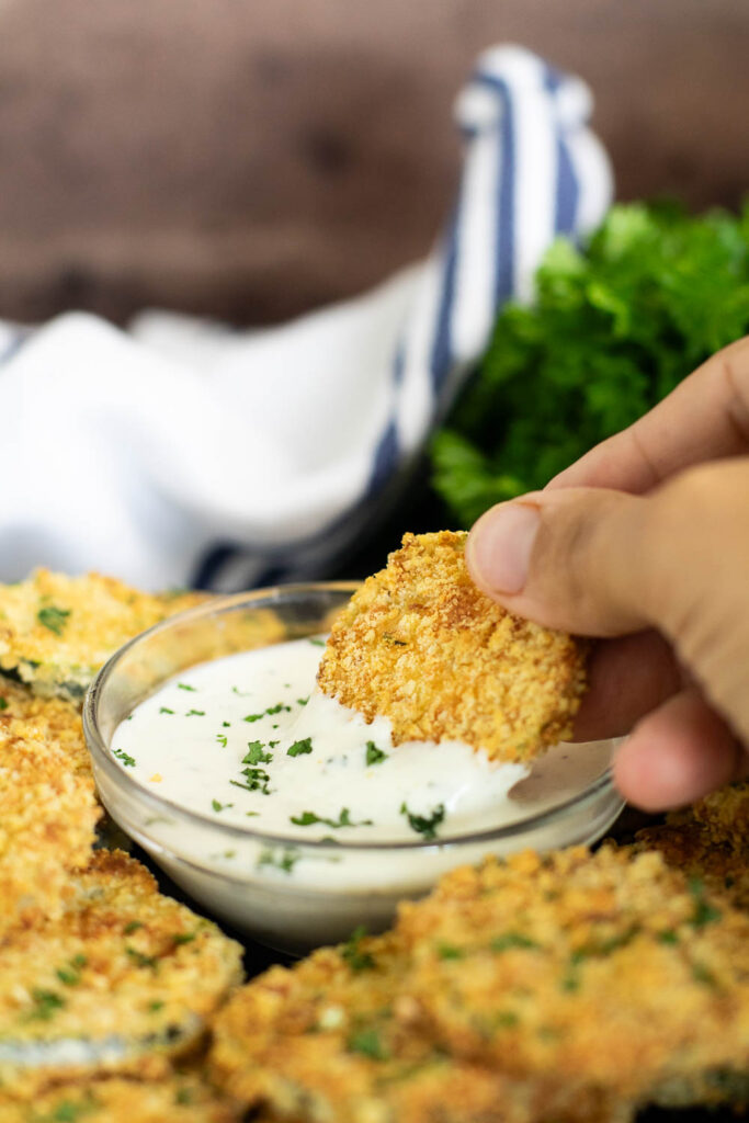 Breaded Air Fryer zucchini chip being dipped into a small glass bowl of ranch dressing surrounded by more zucchini chips on a small black plate.