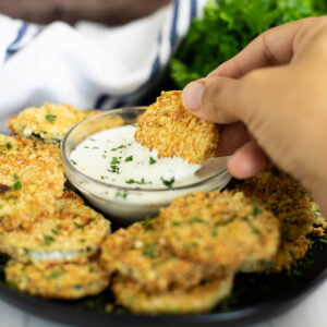 Breaded Air Fryer zucchini chip being dipped into a small glass bowl of ranch dressing surrounded by more zucchini chips on a small black plate.