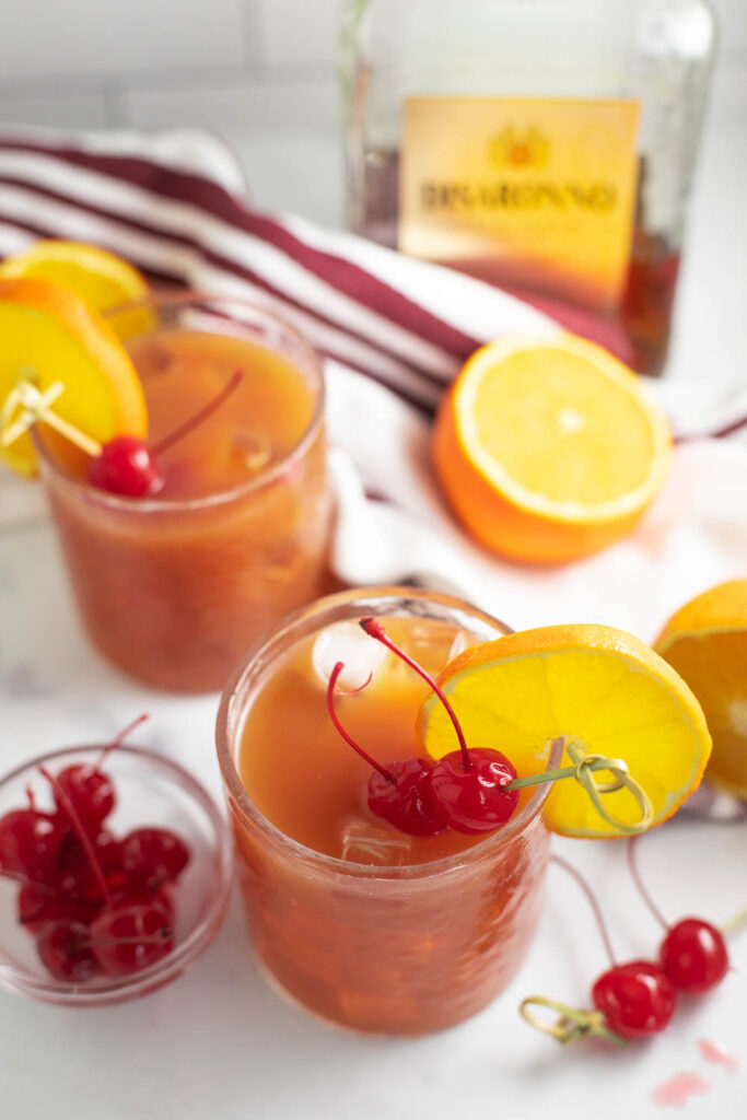 Overhead view of an Alabama slammer drink in a small glass garnished with an orange slice and two maraschino cherries on a toothpick with a second drink, a small bowl of cherries, and a slice orange around it.