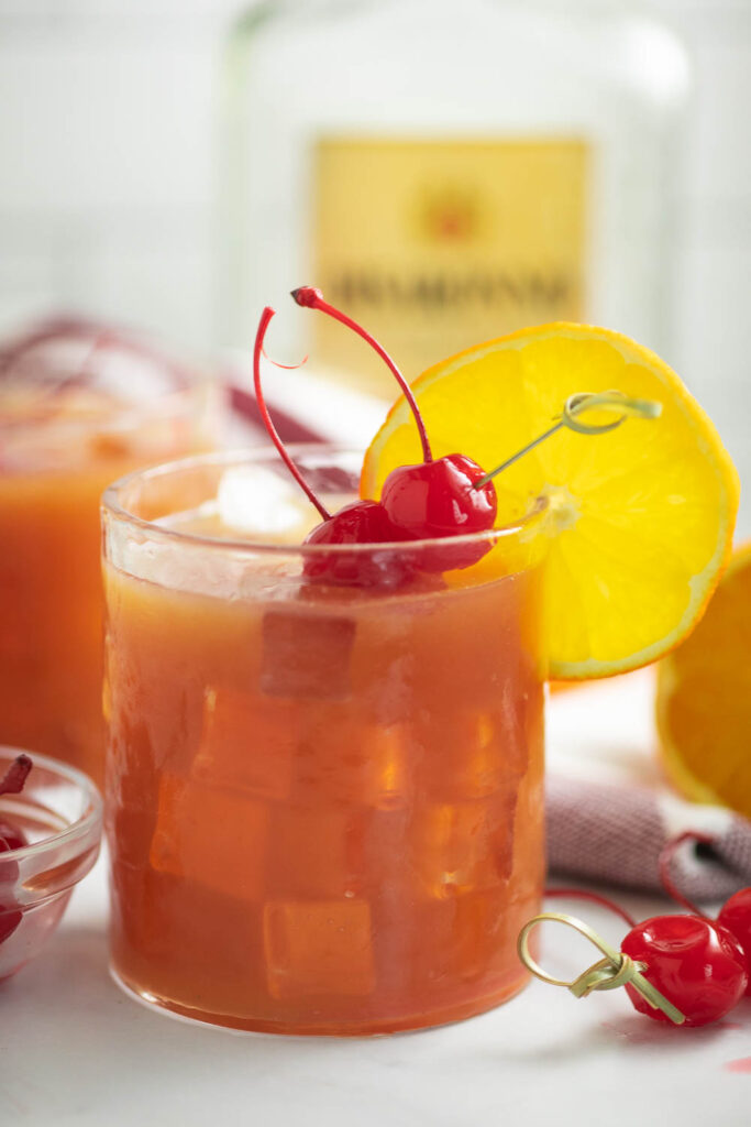 Side view of an Alabama slammer drink in a small glass with an orange slice on the rim and two maraschino cherries on a toothpick in the drink with another drink behind.