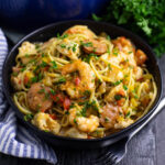 A bowl of pastalaya with shrimp, sausage, and chicken topped with fresh chopped parsley next to a fork, linen napkin, and a bunch of parsley.