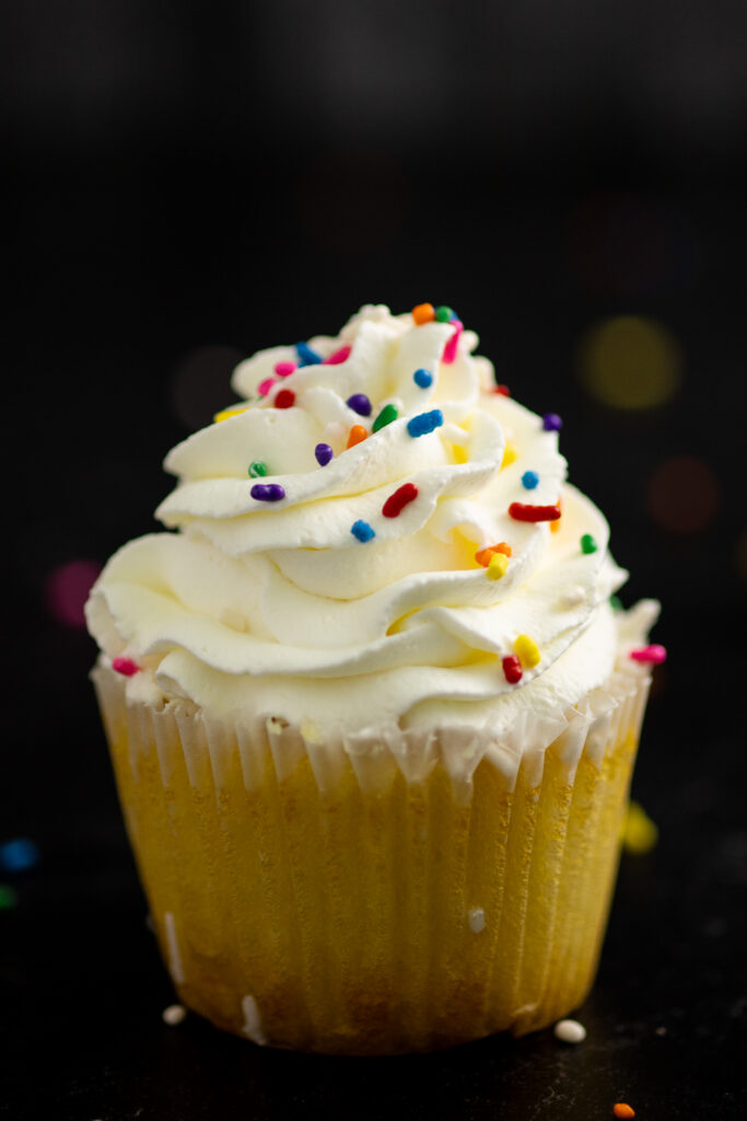 A vanilla cupcake topped with piped stabilized whipped cream and rainbow sprinkles on a black background.