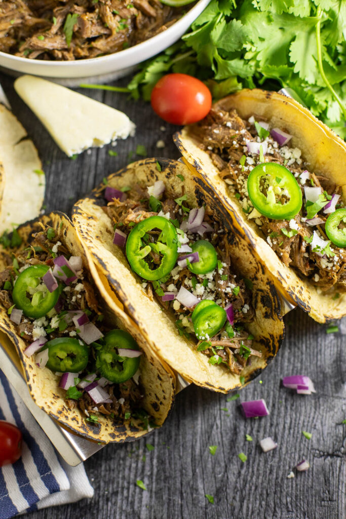Overhead view of three instant pot barbacoa tacos topped with red onions, jalapeno slices, and fresh chopped cilantro surrounded by cotija cheese, a linen napkin, and a bunch of fresh cilantro.