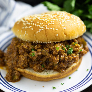 Instant Pot sloppy joe served on a sesame seed bun on a small white plate with a bunch of fresh parsley behind it.