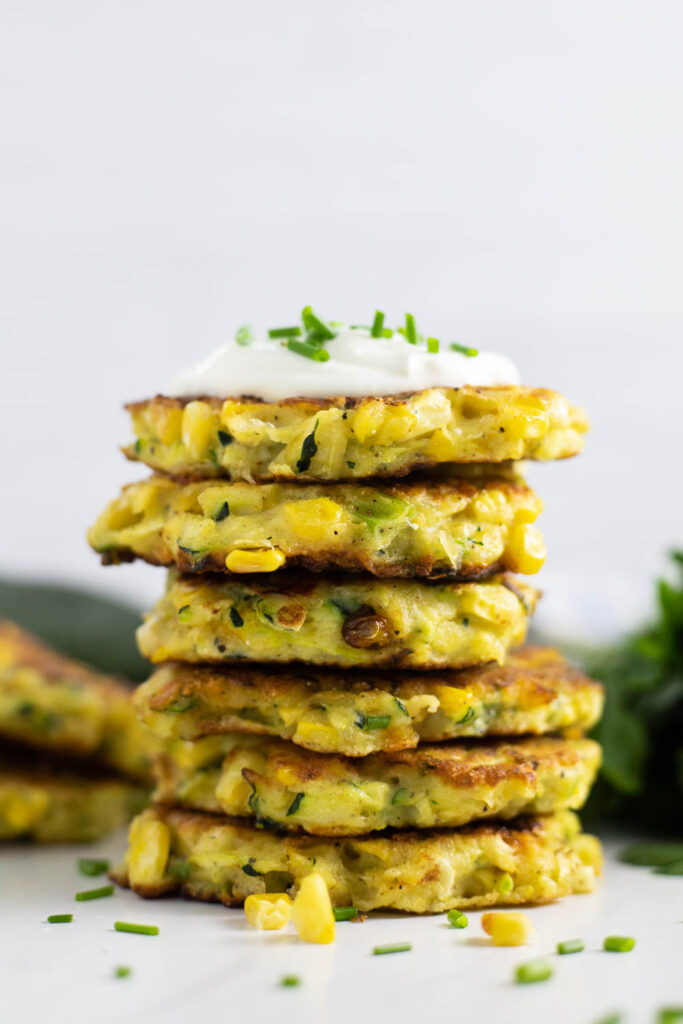 Side view of six zucchini and corn fritters stacked on top of each other topped with sour cream and fresh chives with a bunch of fresh parsley and more fritters behind the stack.