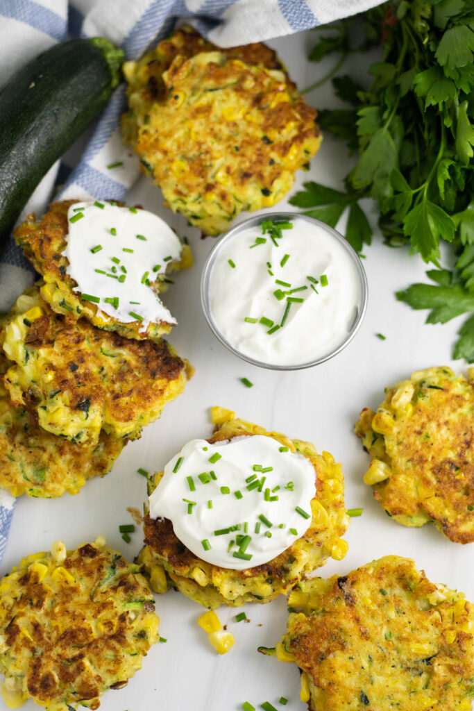 Overhead view of zucchini and corn fritters scattered on a white marble background with a fresh zucchini, bunch of parsley, and cup of sour cream topped with fresh chives. The center fritter is also topped with sour cream and chives.