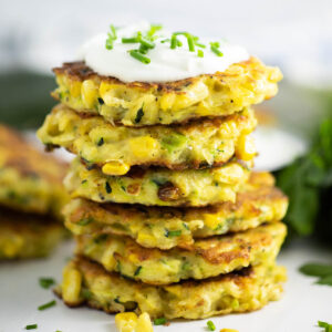 Six zucchini and corn fritters stacked on top of each other with the top fritter topped with sour cream and fresh chives.