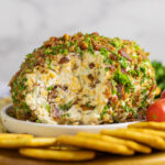 Side view of a bacon ranch cheese ball coated in crumbled bacon and fresh chopped parsley with a section missing to expose the creamy interior, sitting on a plate surrounded by crackers and cherry tomatoes.