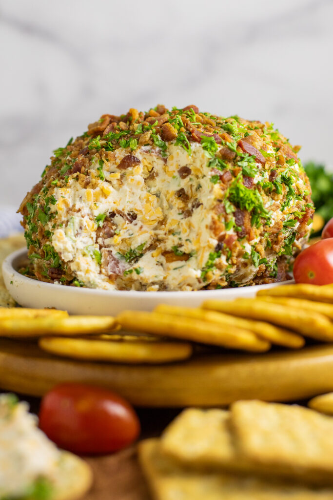 Side view of a bacon ranch cheese ball coated in crumbled bacon and fresh chopped parsley with a section missing to expose the creamy interior, sitting on a plate surrounded by crackers and cherry tomatoes.