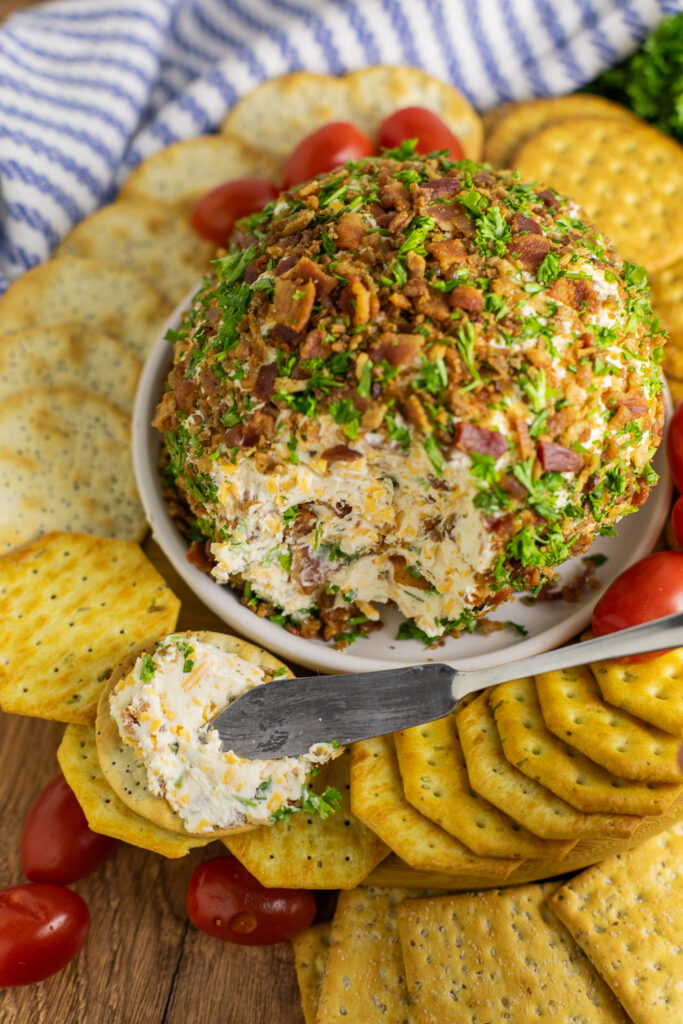 Overhead view of a bacon ranch cheeseball surrounded by crackers, cherry tomatoes, and a linen napkin with one cracker getting the cheeseball smeared onto it in front.