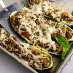 A metal spatula lifting a bruschetta zucchini boat off of a baking sheet next to a sprig of fresh basil.