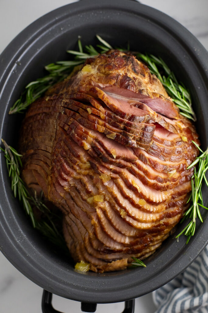 An overhead view of a spiral cut crock pot ham still in the slow cooker surrounded by fresh rosemary sprigs.