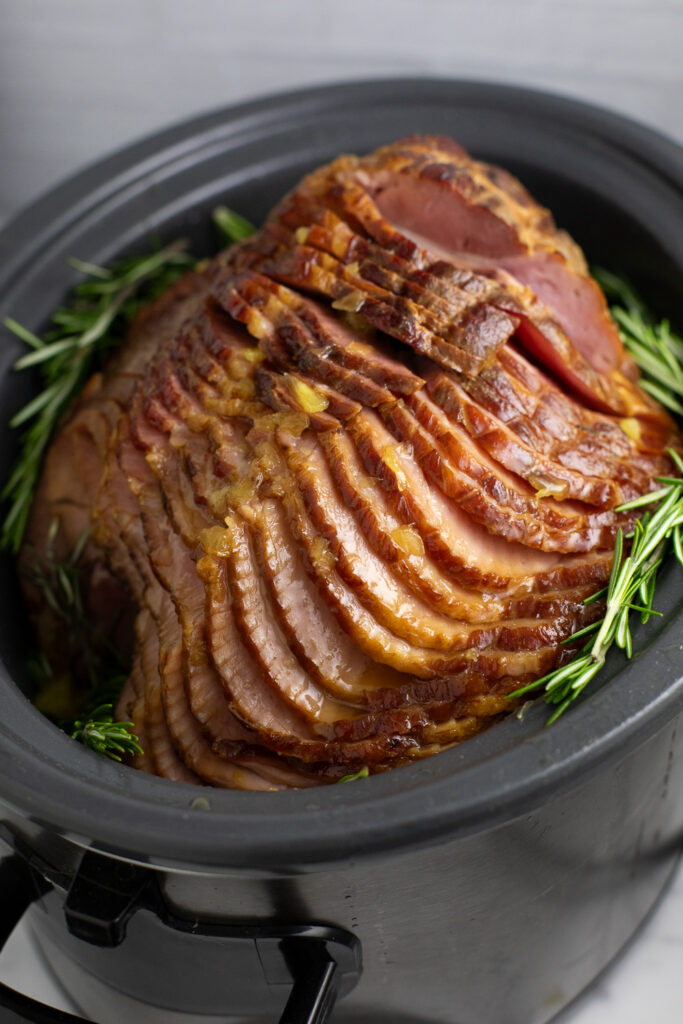A side view of a spiral cut crock pot ham still in the slow cooker surrounded by fresh rosemary sprigs.