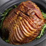 A side view of a spiral cut crock pot ham still in the slow cooker surrounded by fresh rosemary sprigs.
